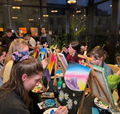 Des participants concentrés en train de peindre des paysages colorés lors d’un atelier de peinture à Paris, dans un cadre lumineux avec vue sur la ville.