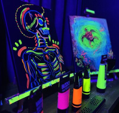 Toiles peintes avec de la peinture fluorescente sous lumière UV, représentant un squelette coloré et une tortue, exposées lors d’un atelier peinture dans le noir à Paris.