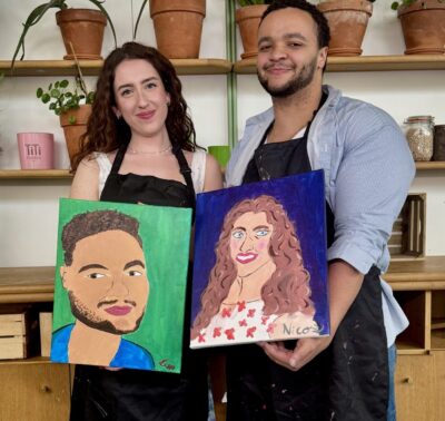 Un couple souriant tenant leurs portraits mutuels peints lors de l’atelier Paint Your Partner à Paris, dans un cadre végétal et chaleureux.