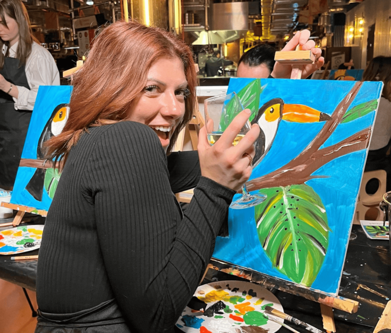Femme souriante tenant un verre de vin devant sa toile représentant un toucan lors d’un atelier de peinture insolite à Paris